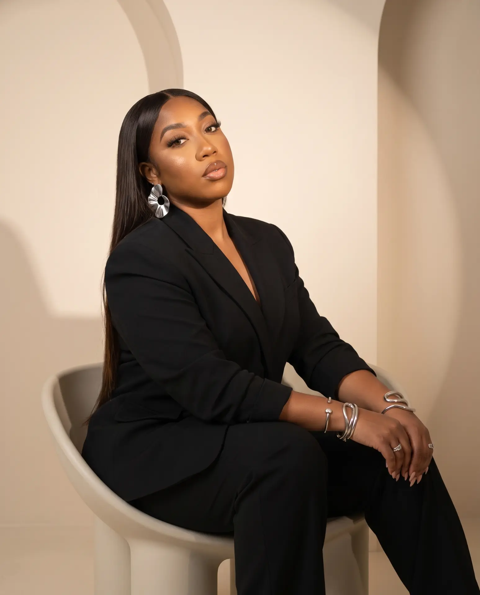 Kendah Parris seated in a tailored black suit with silver jewelry, photographed in a warm studio set.